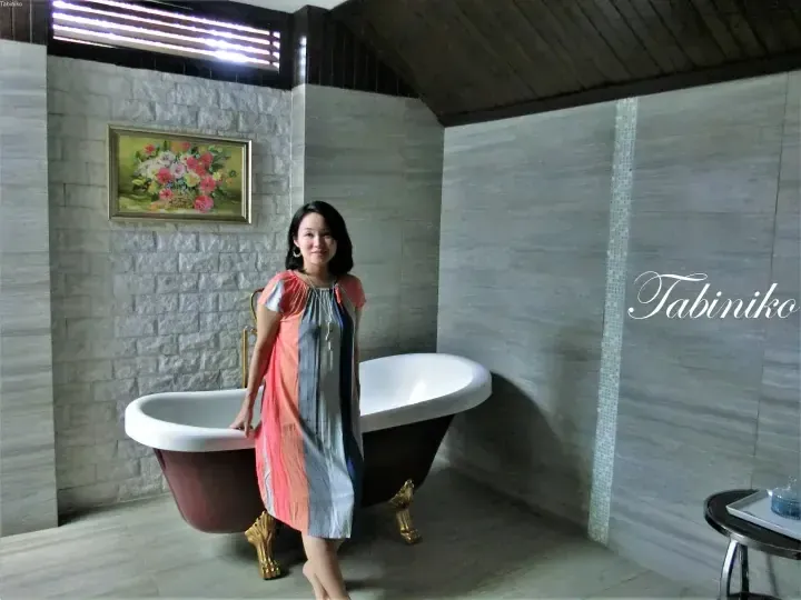 Woman standing beside a freestanding bathtub in a modern bathroom with stone walls and a floral painting.