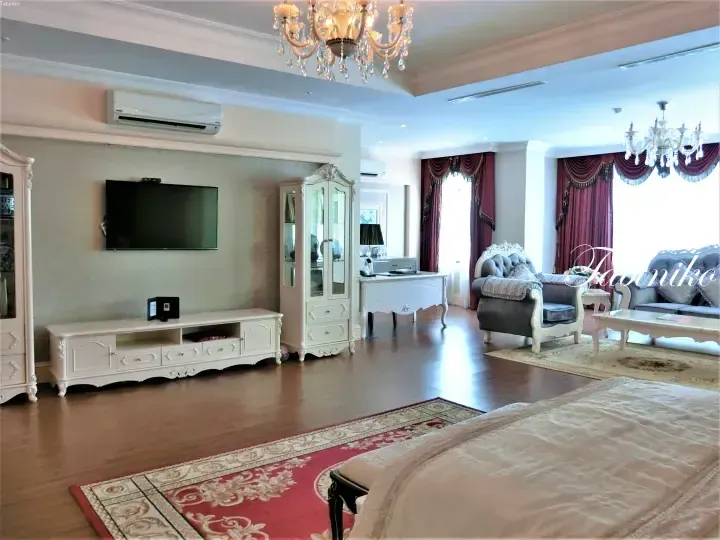 Elegant suite interior featuring a living area with grey sofa, white furniture, and crystal chandeliers, alongside a bedroom with a red patterned rug.