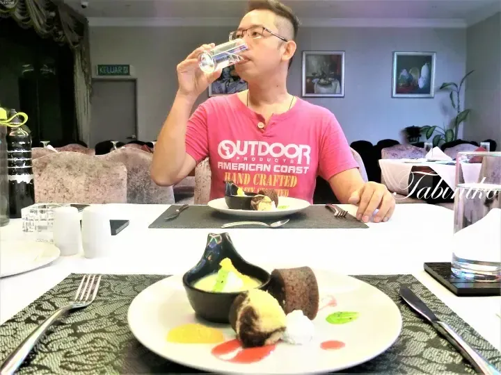 A man in a pink shirt drinks water while dining at a table with dessert plates in a restaurant.