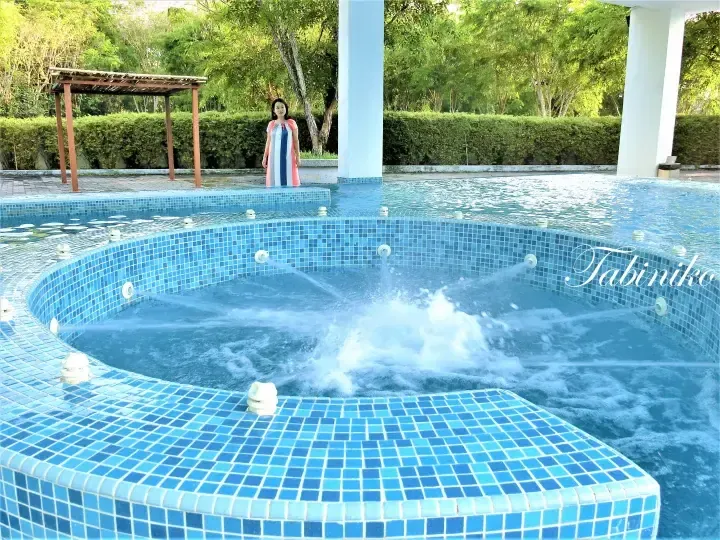 Woman standing by a blue tiled jacuzzi pool with water jets at a resort surrounded by lush greenery.