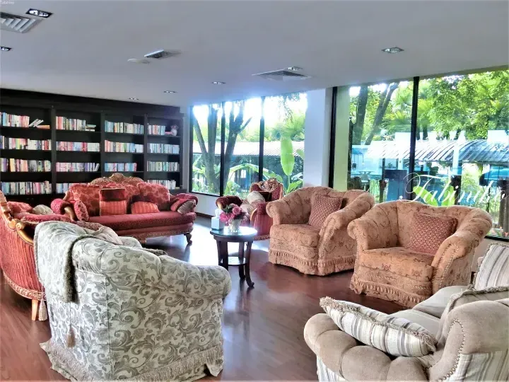 Luxurious lounge with plush sofas and a bookshelf, featuring large windows overlooking tropical greenery.