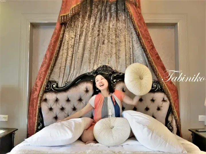 A smiling woman sits on a luxurious bed with a tufted headboard and draped curtains in a hotel room.
