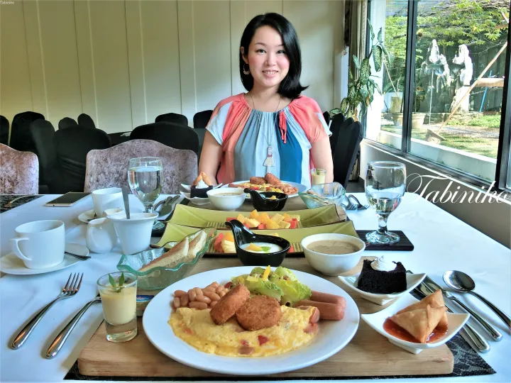 庭園の眺めが楽しめるリゾートで、オムレツやソーセージが並ぶ朝食テーブルに座る女性。