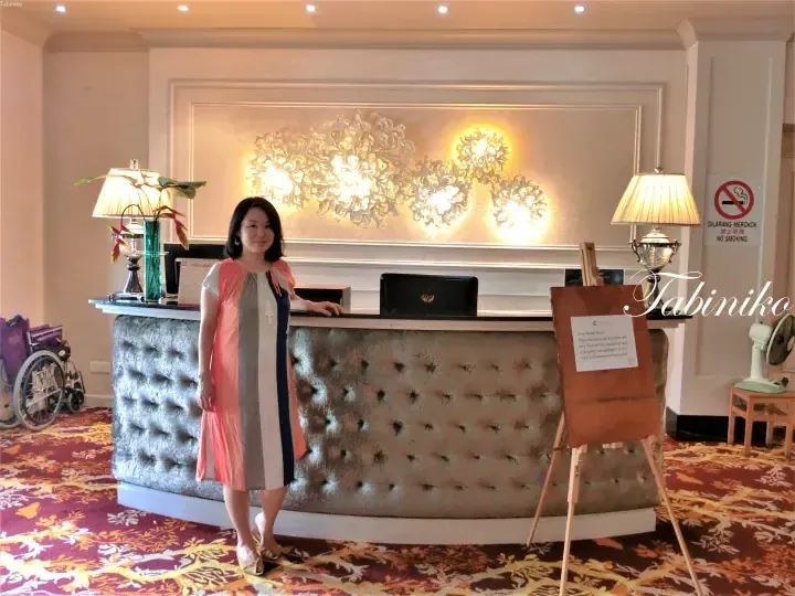 Woman standing at a hotel reception desk with a tufted front, floral wall art, and a notice board in the lobby.