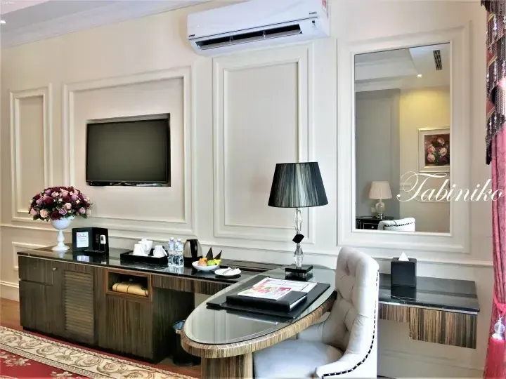 Elegant hotel suite interior featuring a glass desk, floral arrangement, and wall-mounted TV in a cream-colored room.