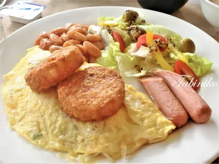 A hearty breakfast plate featuring a fluffy omelette topped with hash browns, baked beans, sausages, and a fresh salad with tomatoes and olives.
