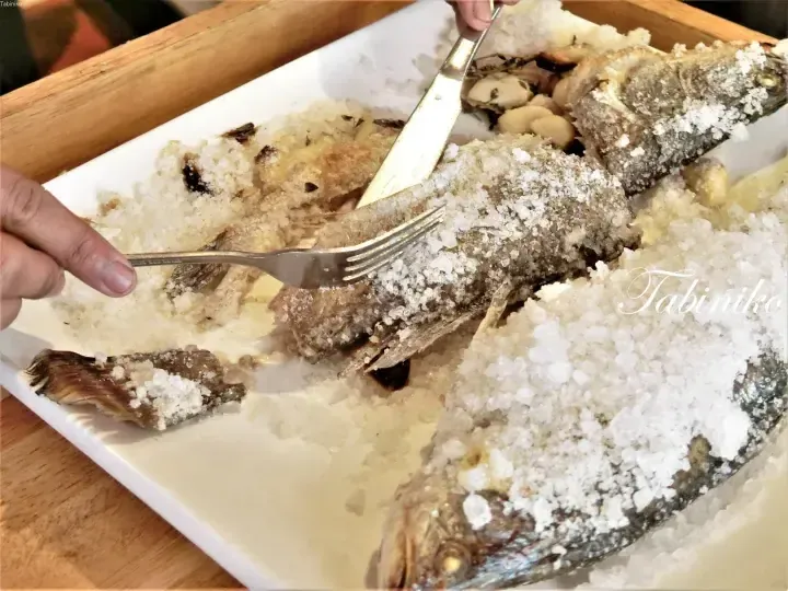 Whole fish baked in a salt crust being broken open with a knife and fork on a white rectangular plate.