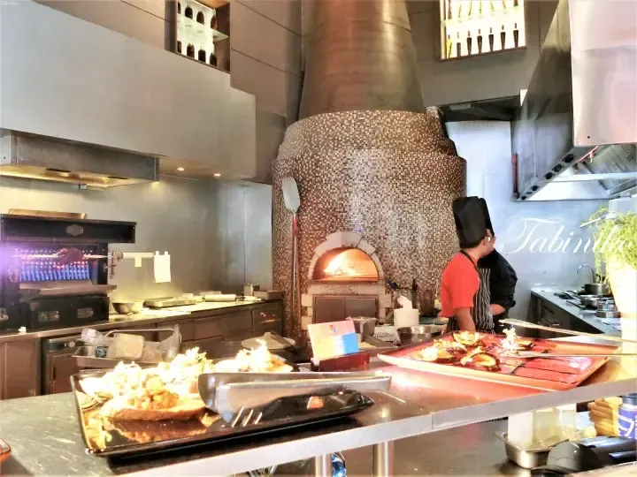 Chef preparing food in a modern kitchen featuring a large mosaic-tiled wood-fired oven and stainless steel appliances.