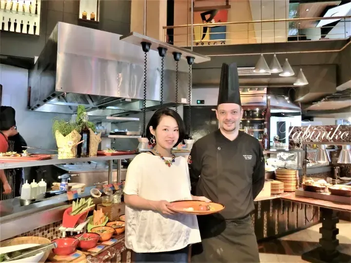 A woman and a chef pose together at a buffet station in a modern restaurant kitchen.