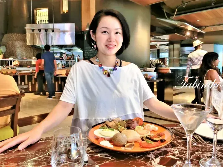 Smiling woman in a white top and colorful necklace sits at a marble table with a plate of assorted buffet food and drinks.
