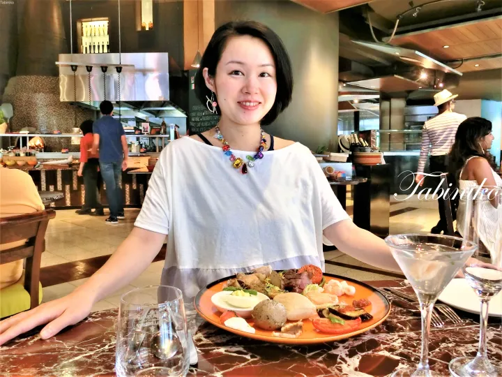 白いトップスとカラフルなネックレスを着た女性が、マラールのテーブルでビュッフェ料理と飲み物を前に微笑んでいる。