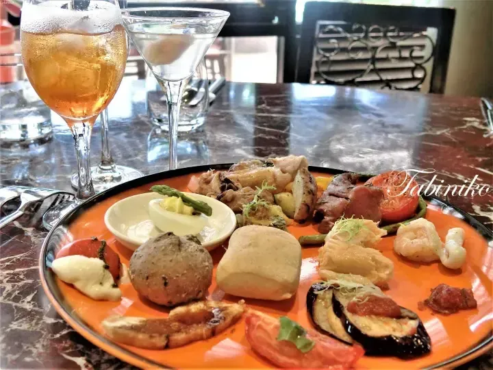 A vibrant orange platter featuring assorted appetizers like bread rolls, shrimp, and eggplant alongside a glass of sparkling drink.