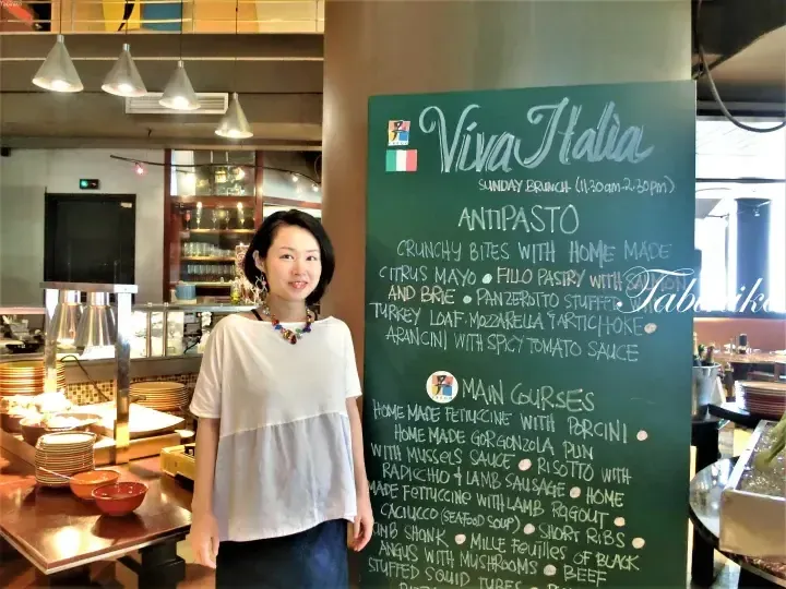 A woman stands beside a chalkboard menu for Viva Italia Sunday Brunch featuring antipasto and main courses.