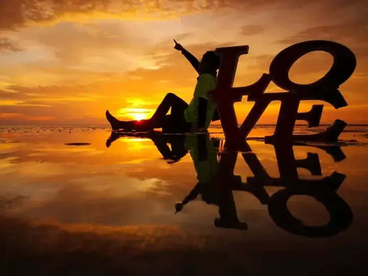 Silhouette of a person sitting on a beach at sunset next to a large LOVE sign with reflection in wet sand.