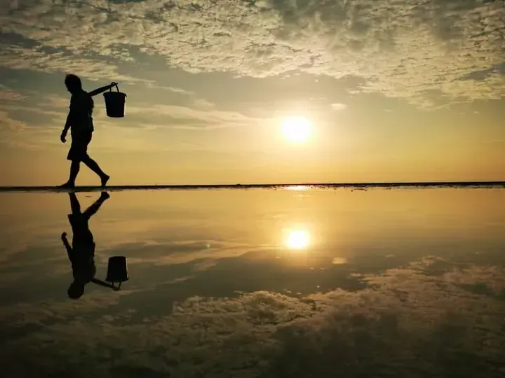 Silhouette of a person carrying a bucket walking on a reflective salt flat at sunset.