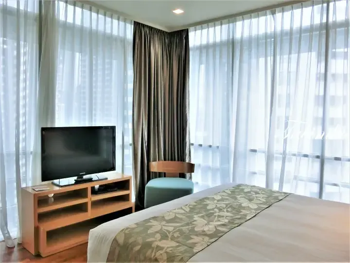 Bright hotel room with a bed, flat-screen TV, and large windows covered by sheer and dark curtains.
