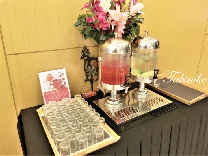 花とAir Sirap Roseの看板が添えられたテーブルに並ぶ、赤と透明の飲料が入ったディスペンサー。