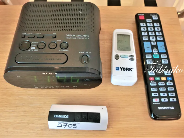 木製テーブルの上に置かれたソニーのドリームマシーン時計ラジオ、ヨークのエアコンリモコン、サムスンのテレビリモコン、トマトのワイヤレスドアベル。