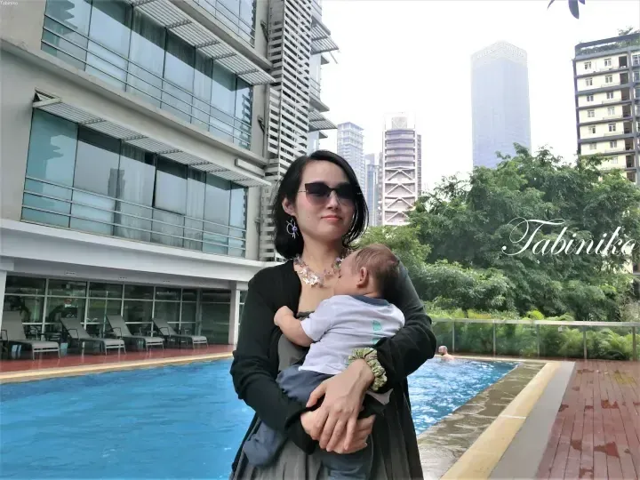 Woman holding a baby by a poolside with Pavilion Kuala Lumpur visible in the background.