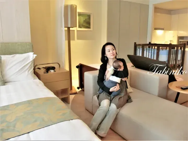Woman holding baby on sofa in modern hotel suite with bed and crib.