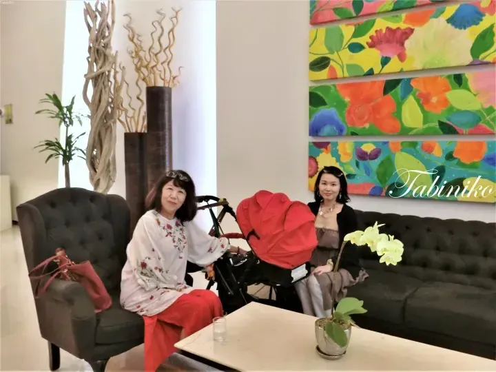 Two women and a red stroller relax in a bright lobby with floral wall art and tall decorative branches.