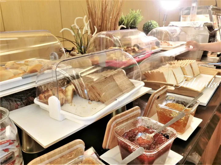 ホテルの朝食ビュッフェで、透明なカバーの下に並べられたパンやペストリー、ジャムの盛り合わせ。