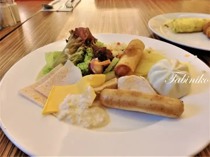 Buffet plate with spring rolls, sausages, chicken, cheese, salad, and steamed buns at a Malaysian hotel breakfast.