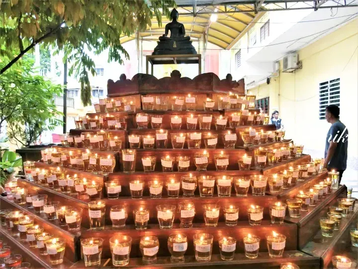 Rows of lit oil lamps on tiered wooden shelves beneath a Buddha statue at a Malaysian temple.