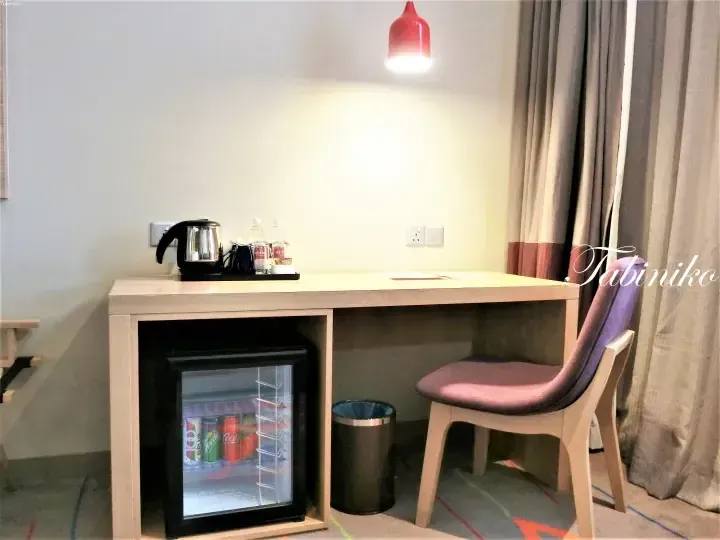 Hotel room desk with kettle, water bottles, mini fridge stocked with drinks, and purple chair under red pendant light