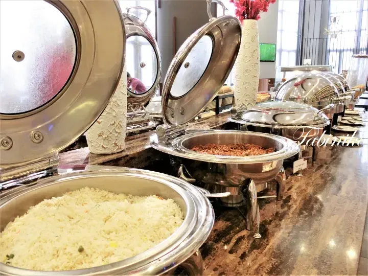 Buffet spread featuring stainless steel chafing dishes with rice and noodles at a hotel dining area.