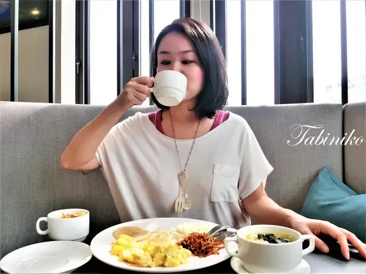 Woman drinking coffee at a breakfast buffet featuring scrambled eggs, rice, and noodles at a Malaysia hotel.