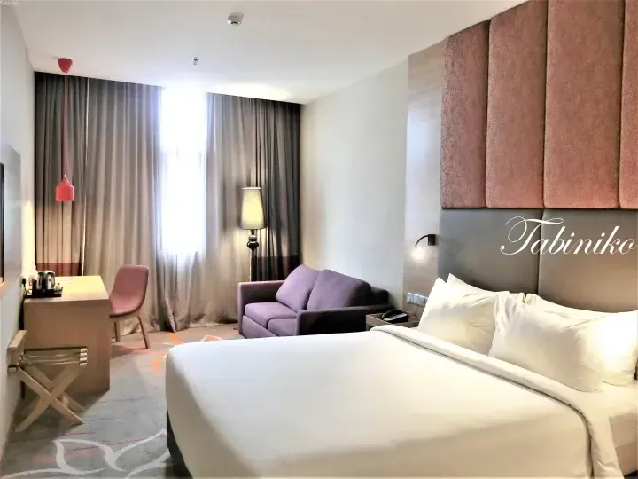 Modern hotel room with a large bed, purple sofa, and desk area featuring pink and grey decor.