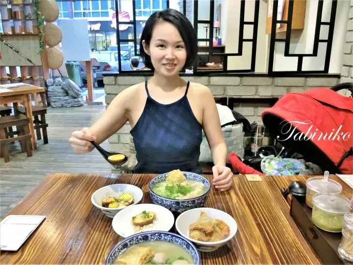 Smiling woman eating soup at a restaurant table with side dishes in Malaysia.