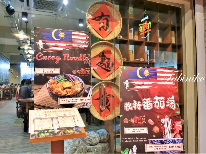 マレーシア国旗と中国書道が飾られたレストランの窓に、カレーヌードルとトマトスープのメニューが展示されている。