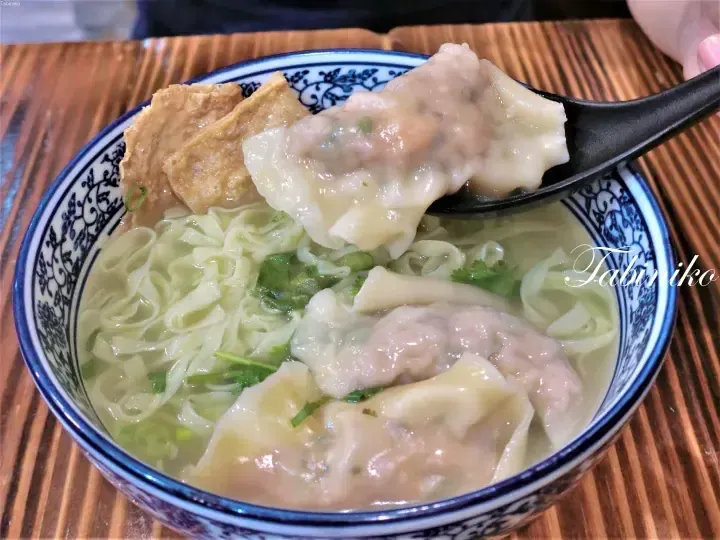A spoon lifts a large wonton from a bowl of wonton noodles with tofu puffs and greens.