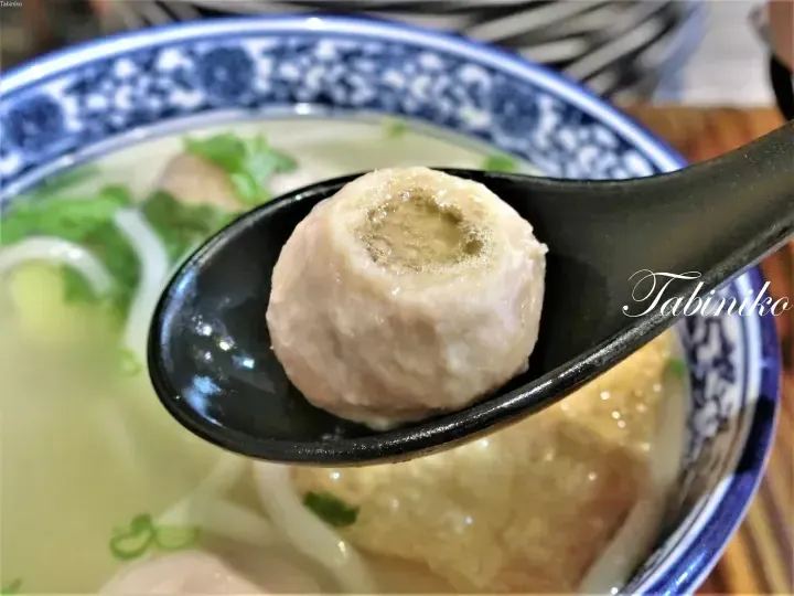 A close-up of a meatball held in a black spoon over a bowl of soup with noodles and herbs.
