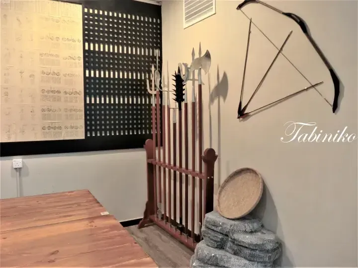 Traditional Malay weapons displayed on a wooden rack alongside a bow and arrow in a room with decorative wall panels.