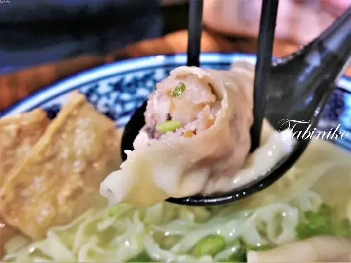 A close-up of a wonton dumpling held in a black spoon, revealing its savory filling, with noodles and fried wontons in a blue and white bowl.