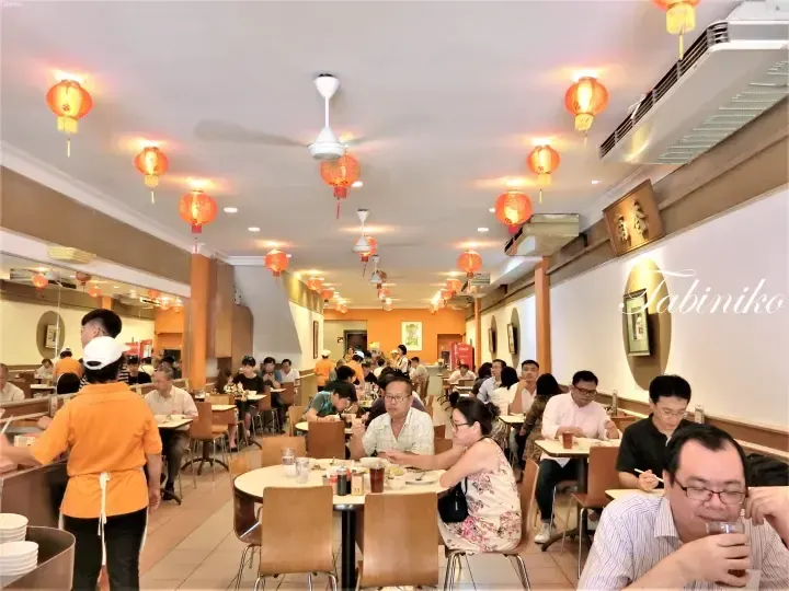 Busy dining hall with red lanterns and ceiling fans, showing patrons eating at tables in a restaurant.