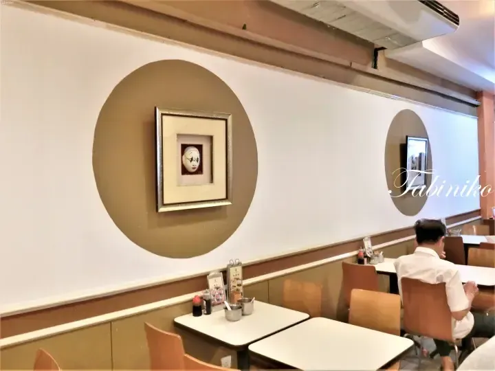 Interior of a restaurant featuring a framed Noh mask on a wall with large circular designs, wooden chairs, and tables.