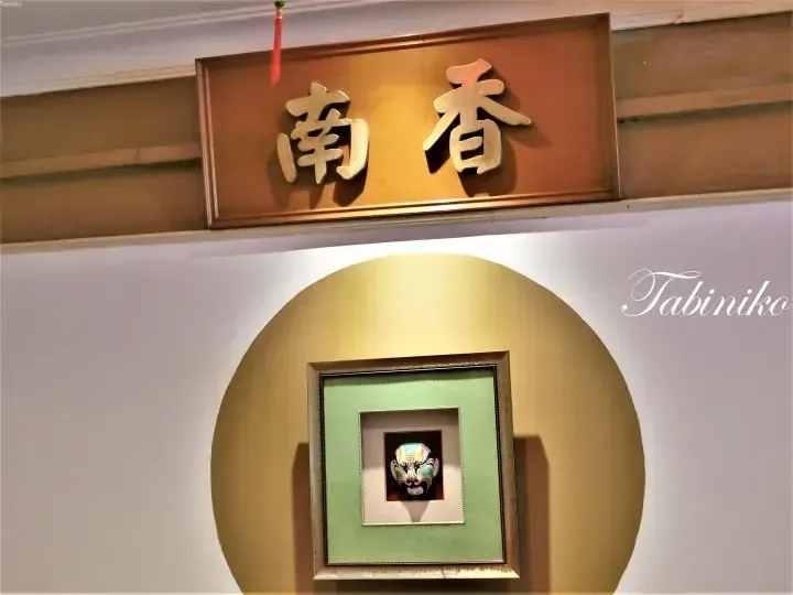 Wooden sign with gold Chinese characters reading Nan Xiang above a framed Peking Opera mask in a circular display.