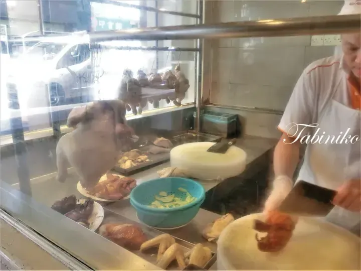 Chef chopping roasted chicken at a food stall with hanging chickens and ingredients visible behind glass.