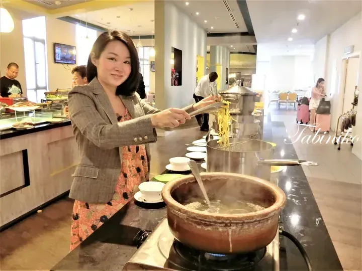 Woman serving noodles at a buffet station in a bright dining area.