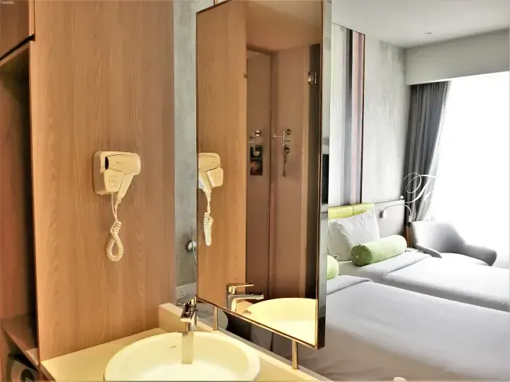 Modern hotel bathroom with a wooden cabinet, wall-mounted hairdryer, and mirror reflecting twin beds with green accents.