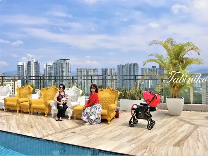 Two women and a baby pose on ornate yellow and white chairs by a rooftop pool with Kuala Lumpur city skyline views.