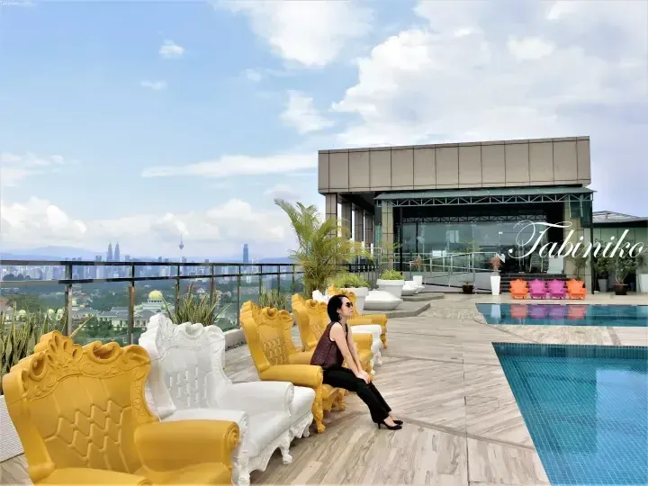 Woman sitting on ornate yellow and white chairs by a rooftop pool overlooking Kuala Lumpur skyline including Petronas Twin Towers.