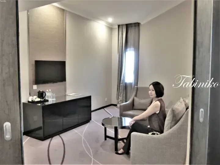 Woman seated in a modern hotel suite living area with a flat-screen TV, minibar, and comfortable seating.