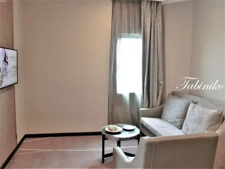 Modern hotel suite living area with grey sofa, fruit plate, and TV displaying Dorsett branding.