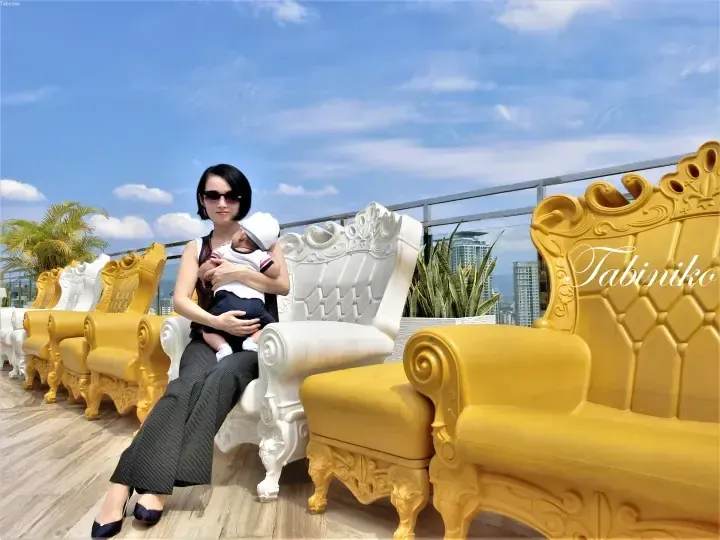 Woman holding a baby on a white throne chair at a rooftop terrace with yellow chairs and city views.