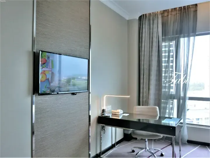 Modern hotel room desk area with a black glass table, white chair, and wall-mounted TV displaying a welcome screen.